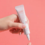 A hand holding Benton Goodbye Redness Centella Spot Cream, applying a small amount onto another hand against a pastel pink background.