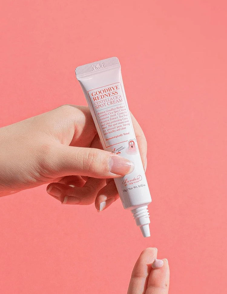 A hand holding Benton Goodbye Redness Centella Spot Cream, applying a small amount onto another hand against a pastel pink background.
