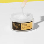 A hand holding a wooden spatula dips into a jar of Cosrx Advanced Snail 92 All in One Cream, showcasing its smooth texture against a yellow background.