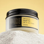A jar of Cosrx Advanced Snail 92 All in One Cream displayed on a textured surface, emphasizing its snail mucin formula for a radiant skin glow.