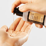 A hand holds a bottle of Cosrx Advanced Snail 96 Mucin Power Essence, dispensing its clear, gel-like texture into the palm below.