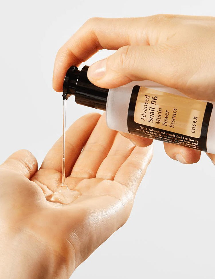 A hand holds a bottle of Cosrx Advanced Snail 96 Mucin Power Essence, dispensing its clear, gel-like texture into the palm below.