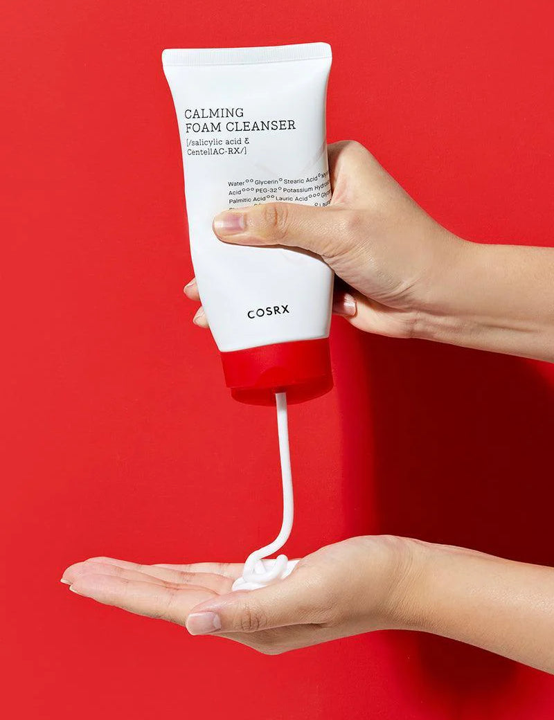 A hand dispenses creamy foam from a white tube of Cosrx Calming Foam Cleanser against a vibrant red background.