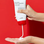 A hand dispenses creamy foam from a white tube of Cosrx Calming Foam Cleanser against a vibrant red background.