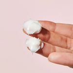 A hand holds two dollops of Cosrx Ceramide Cream, highlighting its creamy texture against a soft pink background.
