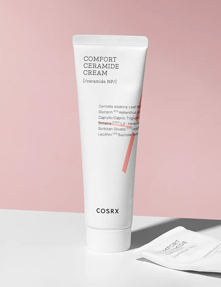 A tube of Cosrx Comfort Ceramide Cream stands on a white surface against a soft pink backdrop, highlighting its minimalist design.