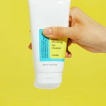 A hand holds a tube of Cosrx Low pH Good Morning Gel Cleanser, pouring gel onto a bright yellow background.