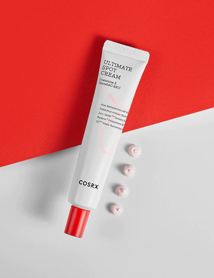 A tube of Cosrx Ultimate Spot Cream with a red cap, placed on a red and white background, alongside small dollops of cream.