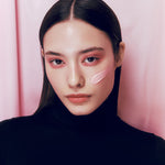 A model with sleek, straight hair wearing a black turtleneck, showcasing smooth, glowing pink cream texture of d'Alba Waterfull Tone-Up Sunscreen on her cheek, standing against a soft pink curtain background.