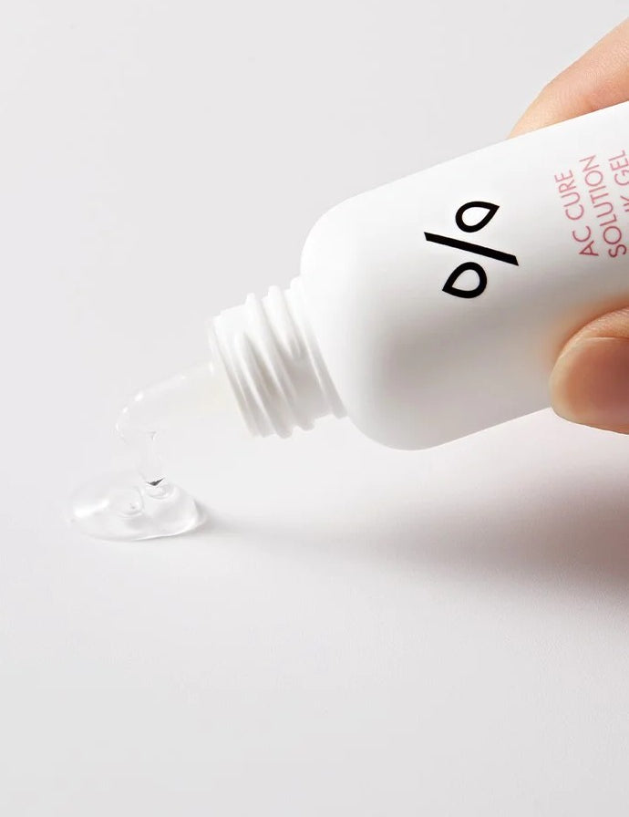 A hand holding a bottle of Dr. Ceuracle AC Cure Solution Pink Gel, with a clear gel droplet spilling onto a plain surface, showcasing its lightweight and soothing texture.