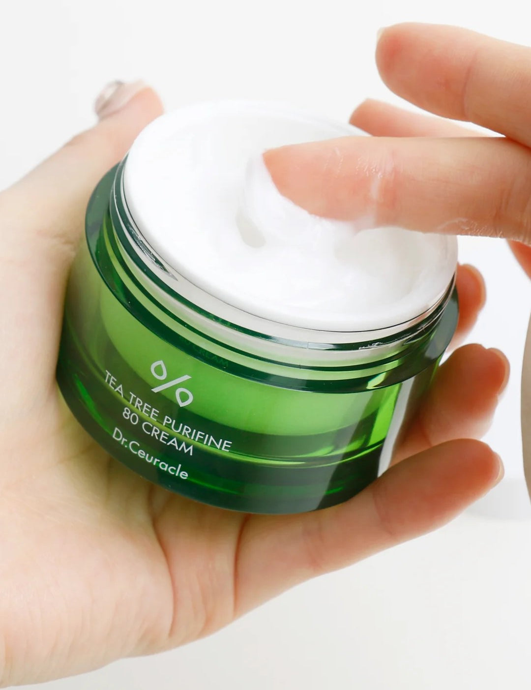 A hand dipping into a green jar of Dr. Ceuracle Tea Tree Purifine 80 Cream, showcasing its creamy texture.