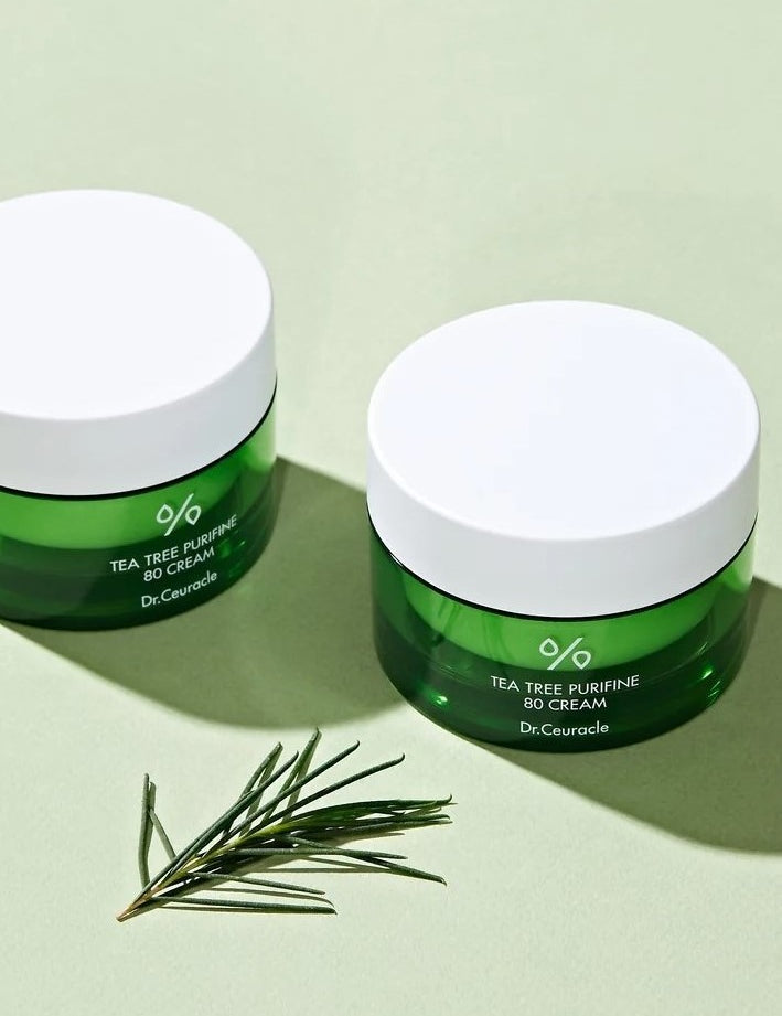 Two green jars of Dr. Ceuracle Tea Tree Purifine 80 Cream on a pale green surface, accompanied by fresh tea tree leaves, highlighting its soothing and clarifying properties.