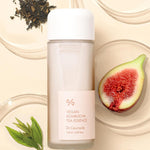 A bottle of Dr. Ceuracle Vegan Kombucha Tea Essence, surrounded by Kombucha Tea leaves, a halved fig, Schisandra chinensis berries, and assorted leaves, set against a cream background.