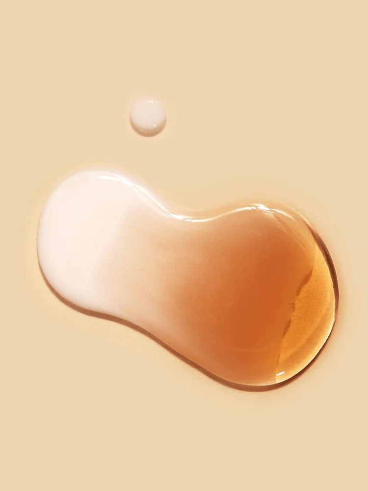 Two droplets of liquid, one creamy white and one amber, rest on a smooth beige surface, showcasing the cream-essence texture of Dr.Ceuracle Vegan Kombucha Tea Essence.