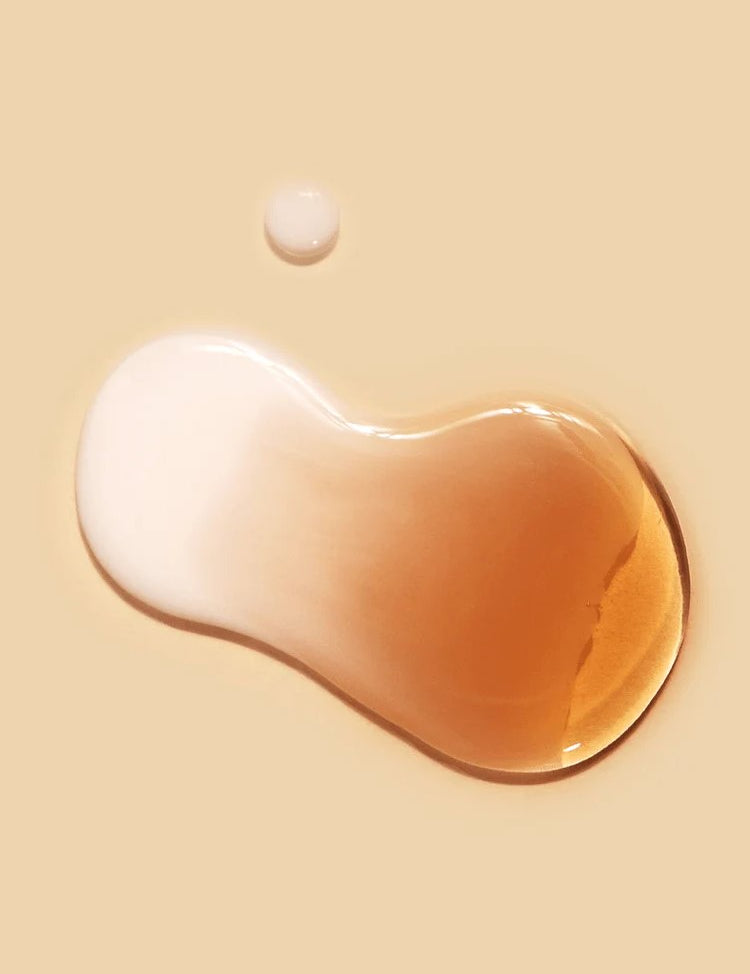 Two droplets of liquid, one creamy white and one amber, rest on a smooth beige surface, showcasing the cream-essence texture of Dr.Ceuracle Vegan Kombucha Tea Essence.