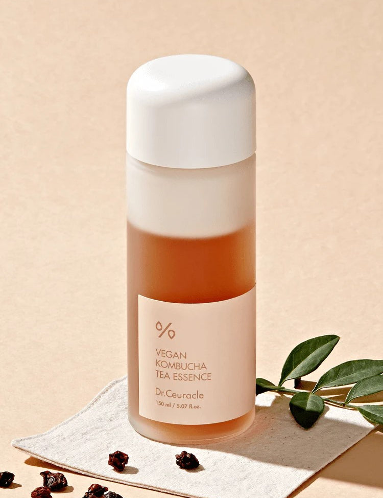 A sleek bottle of Dr.Ceuracle Vegan Kombucha Tea Essence on a neutral background, accompanied by green leaves and dried berries.