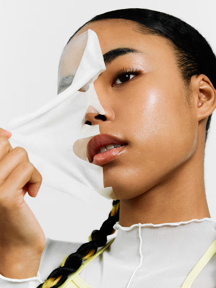 A model with long braided hair holds a Dr. Jart+ Ceramidin Skin Barrier Moisturizing Mask, wearing a stylish gray and yellow outfit against a neutral background.