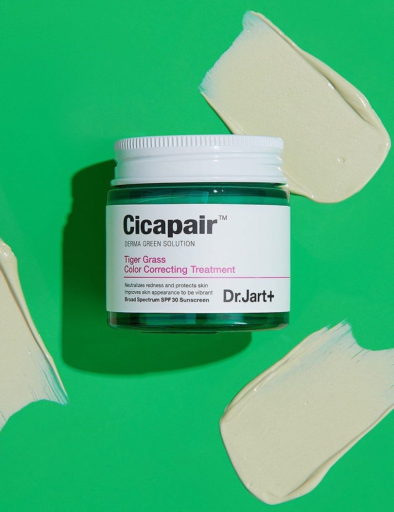 Jar of Dr. Jart+ Cicapair Tiger Grass Color Correcting Treatment on a vibrant green background, with a creamy swatch showcasing its soothing and color-correcting properties.