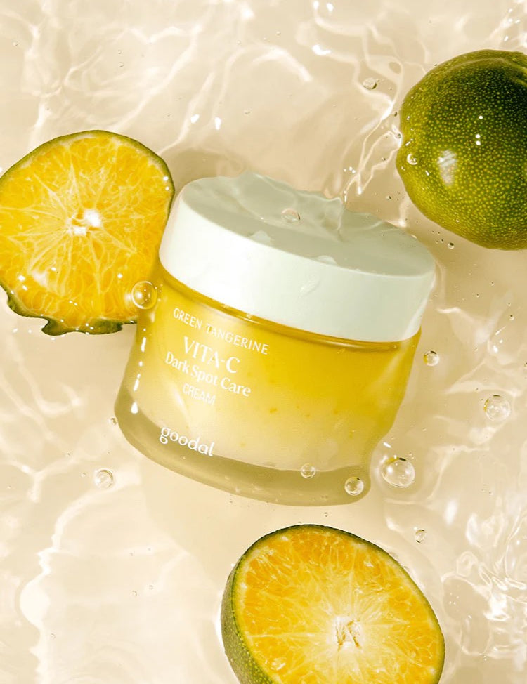 A jar of Goodal Vita-C Dark Spot Care Cream surrounded by fresh green tangerines, its main ingredient, floating against a light, watery background.