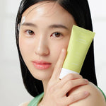 A model with sleek black hair, wearing a green top, holds a tube of Goodal Houttuynia Cordata Calming Moisture Sun Cream against her shoulder in soft lighting.