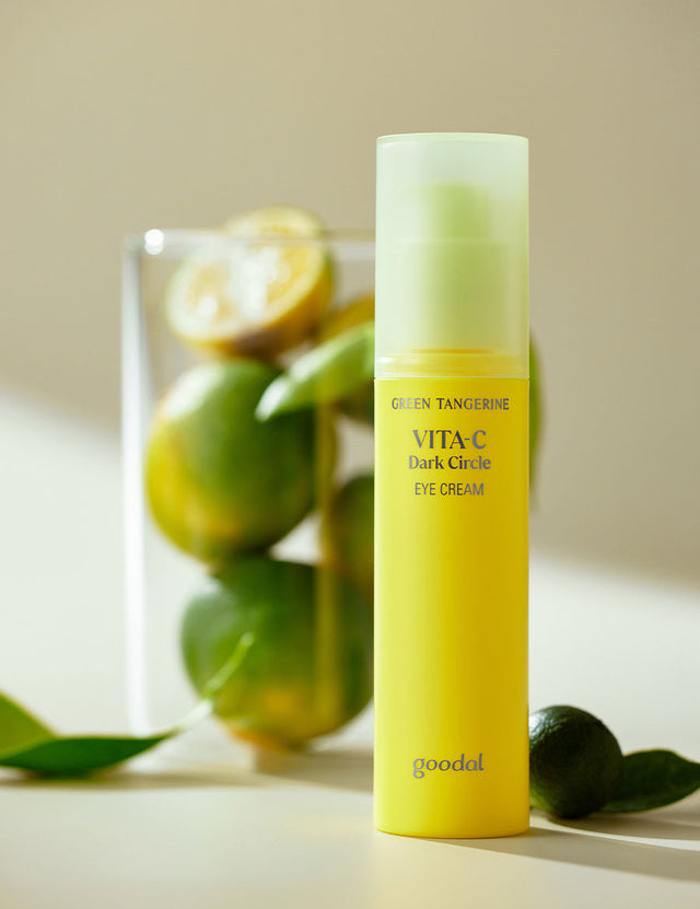 A bright yellow bottle of Goodal Green Tangerine Vita C Dark Circle Eye Cream, set on a soft, light surface with green tangerines in the background, highlighting its main ingredient.