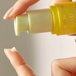 A hand holds a yellow bottle of Goodal Green Tangerine Vita C Dark Circle Eye Cream, with a droplet of cream on the fingertip, showcasing its smooth texture.