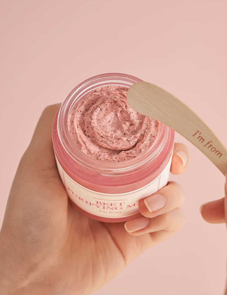 A hand holding a jar of 'I'm From Beet Purifying Mask' pink cream with a wooden spatula hovering above, set against a soft pink background, highlighting its creamy, antioxidant-rich formula.