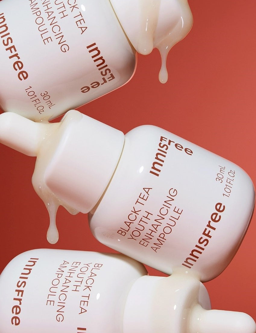 Three Innisfree Black Tea Youth Enhancing Ampoule bottles, white with amber text, dripping essence against a coral background, highlighting its luxurious and rejuvenating formula.