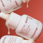 Three Innisfree Black Tea Youth Enhancing Ampoule bottles, white with amber text, dripping essence against a coral background, highlighting its luxurious and rejuvenating formula.