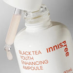 A close-up of Innisfree's Black Tea Youth Enhancing Ampoule with a dropper dispensing a pale liquid, highlighting its sleek white bottle design and rejuvenating formula.