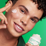 A male model holding a jar of Innisfree Green Tea Seed Hyaluronic Cream against a vibrant green background, showcasing its smooth texture applied to his cheek.