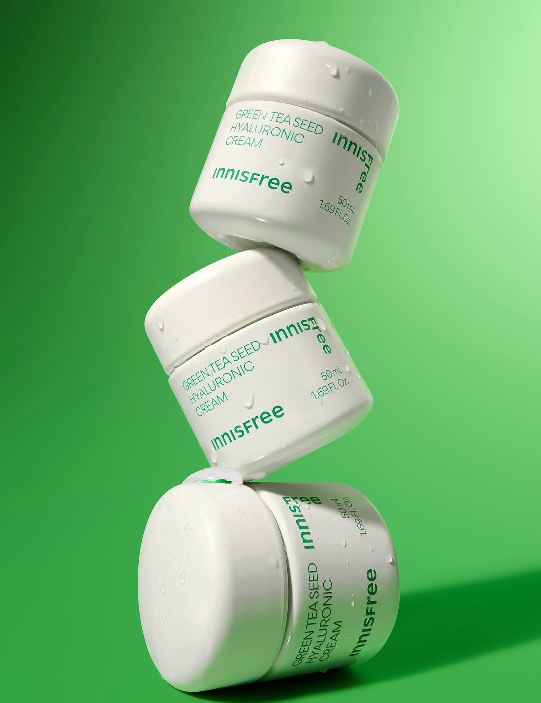 Three Innisfree Green Tea Seed Hyaluronic Cream jars stacked on a vibrant green background, glistening with droplets of water to emphasize hydration and freshness.