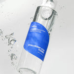 Isntree Hyaluronic Acid Toner with a blue label, surrounded by water droplets on a light backdrop, emphasizing its hydrating properties.