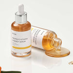 Bottle of iUNIK Propolis Vitamin Synergy Serum with golden liquid spilling from dropper, surrounded by green leaves and sea buckthorn fruit, highlighting nourishing and brightening benefits.