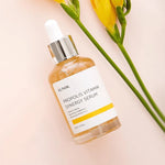 Bottle of iUNIK Propolis Vitamin Synergy Serum on a soft pink background with yellow flowers and green leaves, highlighting its natural and nourishing formula.