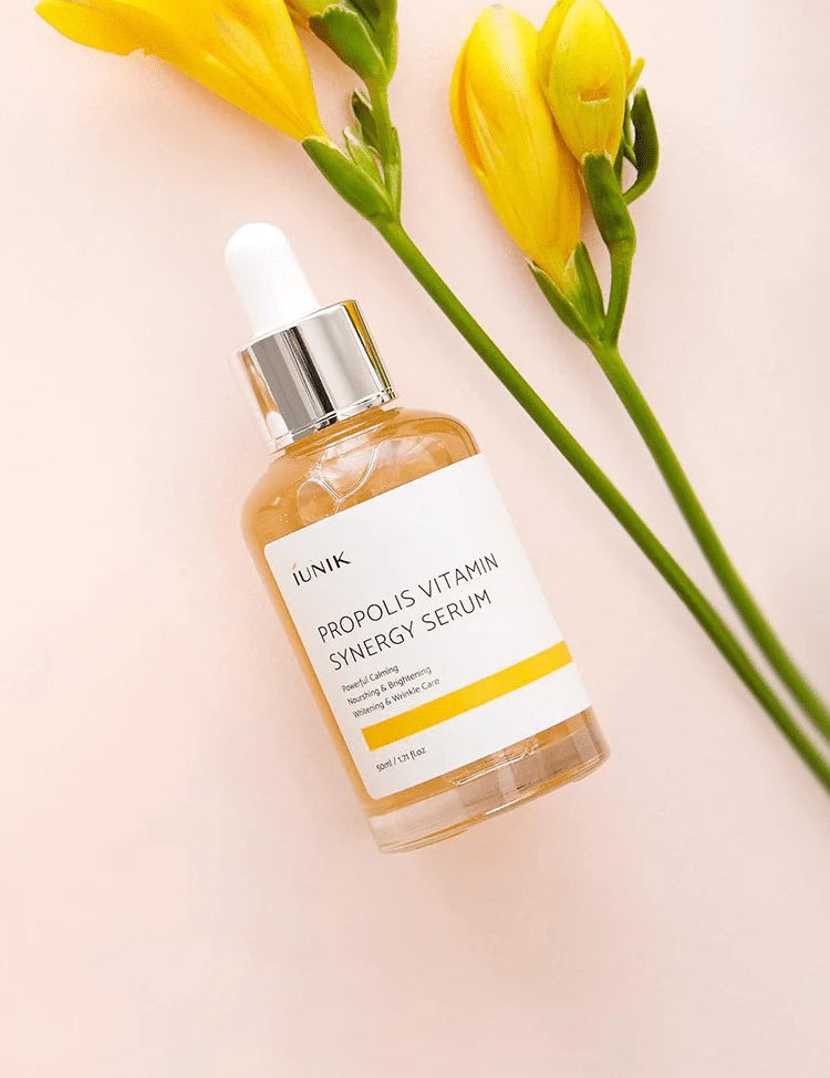 Bottle of iUNIK Propolis Vitamin Synergy Serum on a soft pink background with yellow flowers and green leaves, highlighting its natural and nourishing formula.