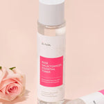 Bottle of iUNIK Rose Galactomyces Essential Toner with pink label next to a delicate pink rose on a soft pastel background, emphasizing gentle hydration effects.