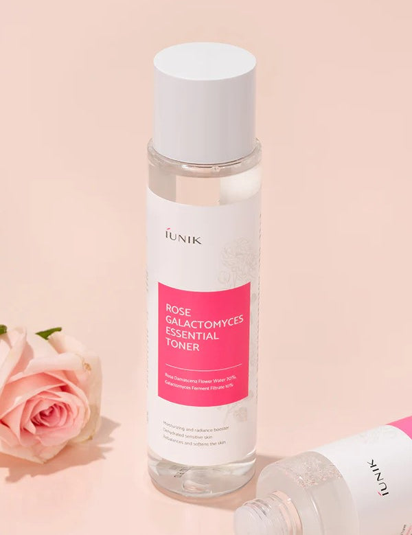 Bottle of iUNIK Rose Galactomyces Essential Toner with pink label next to a delicate pink rose on a soft pastel background, emphasizing gentle hydration effects.
