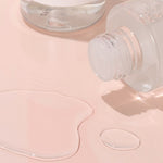 Close-up of iUNIK Rose Galactomyces Essential Toner’s clear, lightweight texture with fluid droplets on a soft pink surface, highlighting its hydrating and soothing properties.