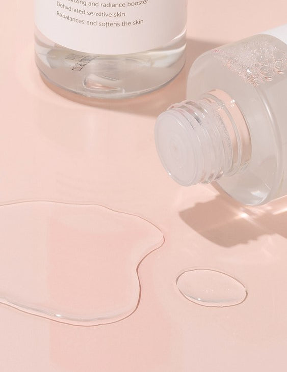 Close-up of iUNIK Rose Galactomyces Essential Toner’s clear, lightweight texture with fluid droplets on a soft pink surface, highlighting its hydrating and soothing properties.