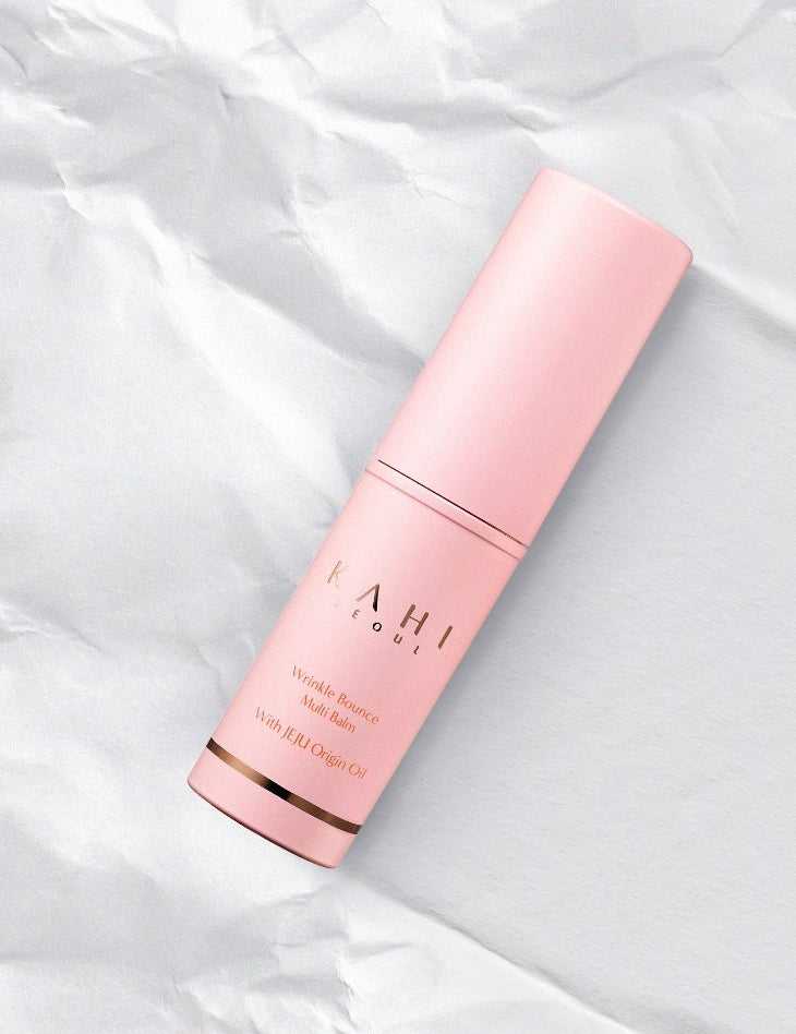 A pink cylinder container of Kahi Wrinkle Bounce Multi Balm with Jeju Origin Oil, placed on a crumpled white paper background.
