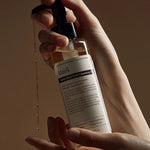 A hand holding Klairs Gentle Black Fresh Cleansing Oil with droplets falling, set against a warm brown background, emphasizing its lightweight texture.