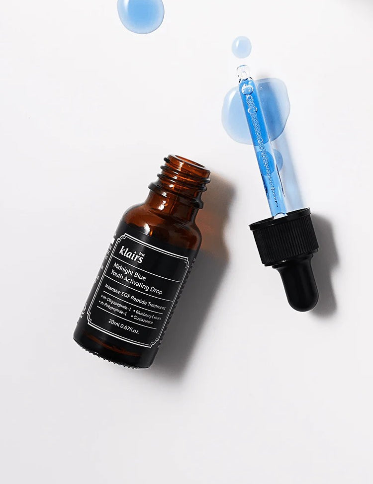 Amber bottle of Klairs Midnight Blue Youth Activating Drop with a dropper, surrounded by vibrant blue liquid droplets on a clean white background, showcasing its lightweight texture and anti-aging properties.