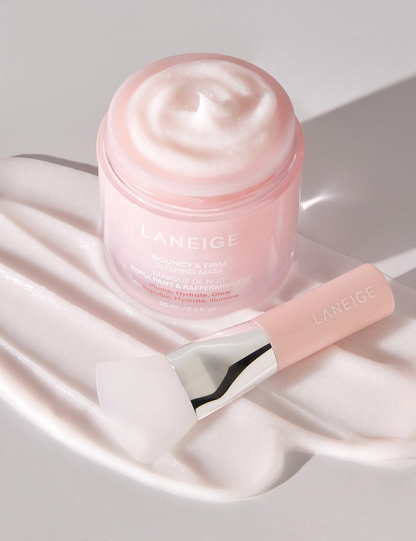 Open jar of Laneige Bouncy & Firm Sleeping Mask with creamy texture and matching applicator on a soft pink background.
