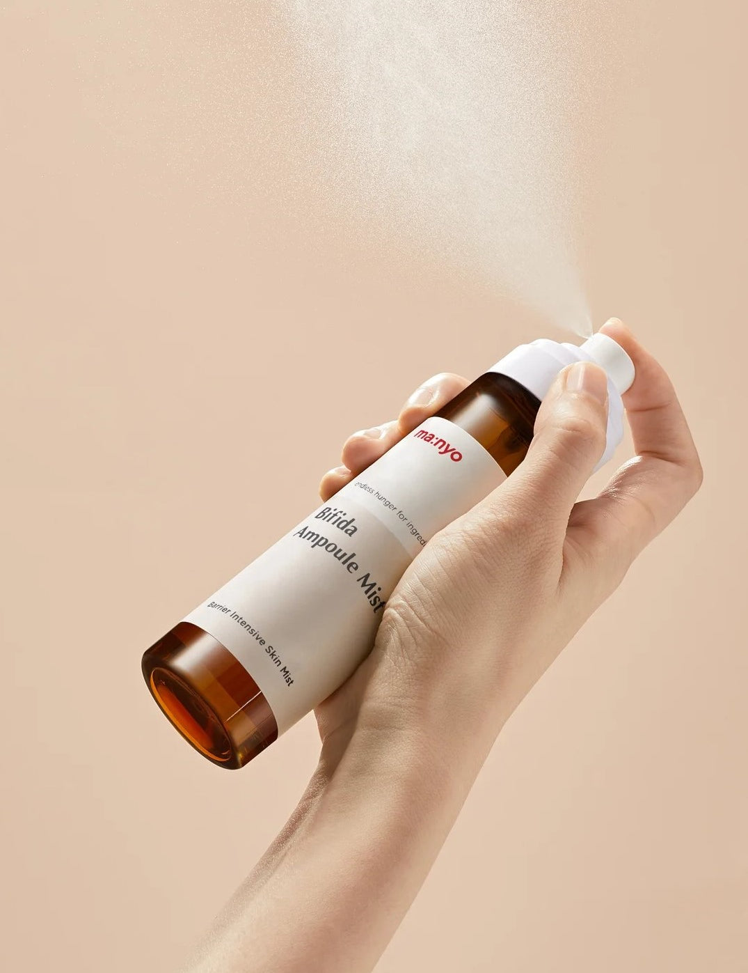 Hand holding a bottle of Manyo Bifida Ampoule Mist releasing a fine mist against a soft beige background, highlighting its lightweight and hydrating formula.