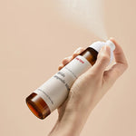 Hand holding a bottle of Manyo Bifida Ampoule Mist releasing a fine mist against a soft beige background, highlighting its lightweight and hydrating formula.