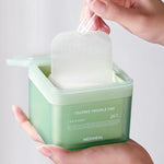 A hand holding a green container of Mediheal Teatree Trouble Pad, lifting a cotton pad out with a clear tool, showcasing ease of use.