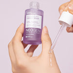 A hand holding a purple bottle of Neogen Dermalogy Real Bakuchiol Firming Serum with a dropper dispensing liquid serum, highlighting its firming and anti-aging properties.