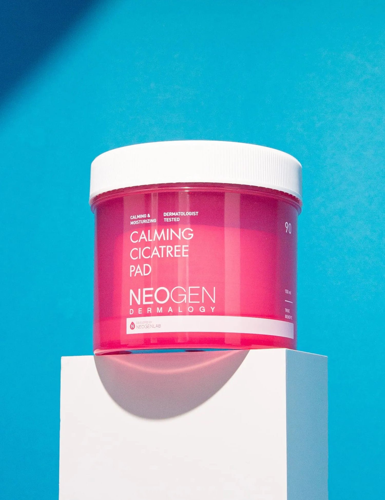 Bright pink jar of Neogen Dermalogy Calming Cicatree Pad displayed on a white pedestal against a vibrant blue background, highlighting its soothing benefits.