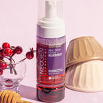 Neogen Real Fresh Foam Blueberry Cleanser bottle displayed alongside fresh blueberries and a honey dipper on a pastel pink backdrop, emphasizing its antioxidant-rich formula and gentle cleansing benefits.